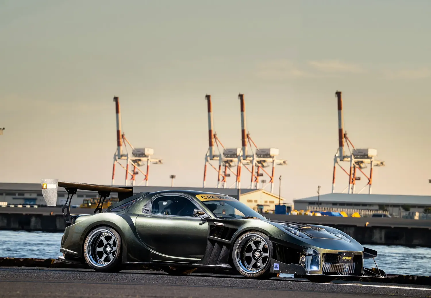港の岸壁に停車したダークグリーンのマツダRX-7 FD3S、ワイドボディと深リム